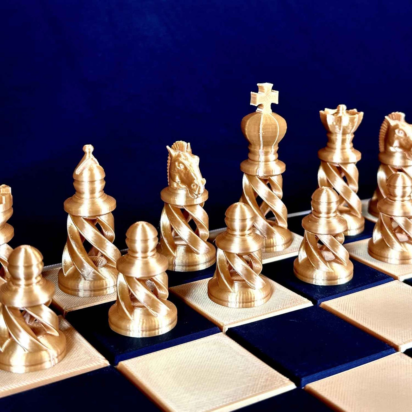 Pièces d’échecs modernes dorées Chess Project alignées sur un échiquier bleu et or, vue de côté mettant en valeur la base torsadée et le design contemporain.